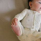 A baby lying on a beige changing pad wearing a cream-colored long-sleeve footless sleeper with a subtle blue and yellow celestial print.