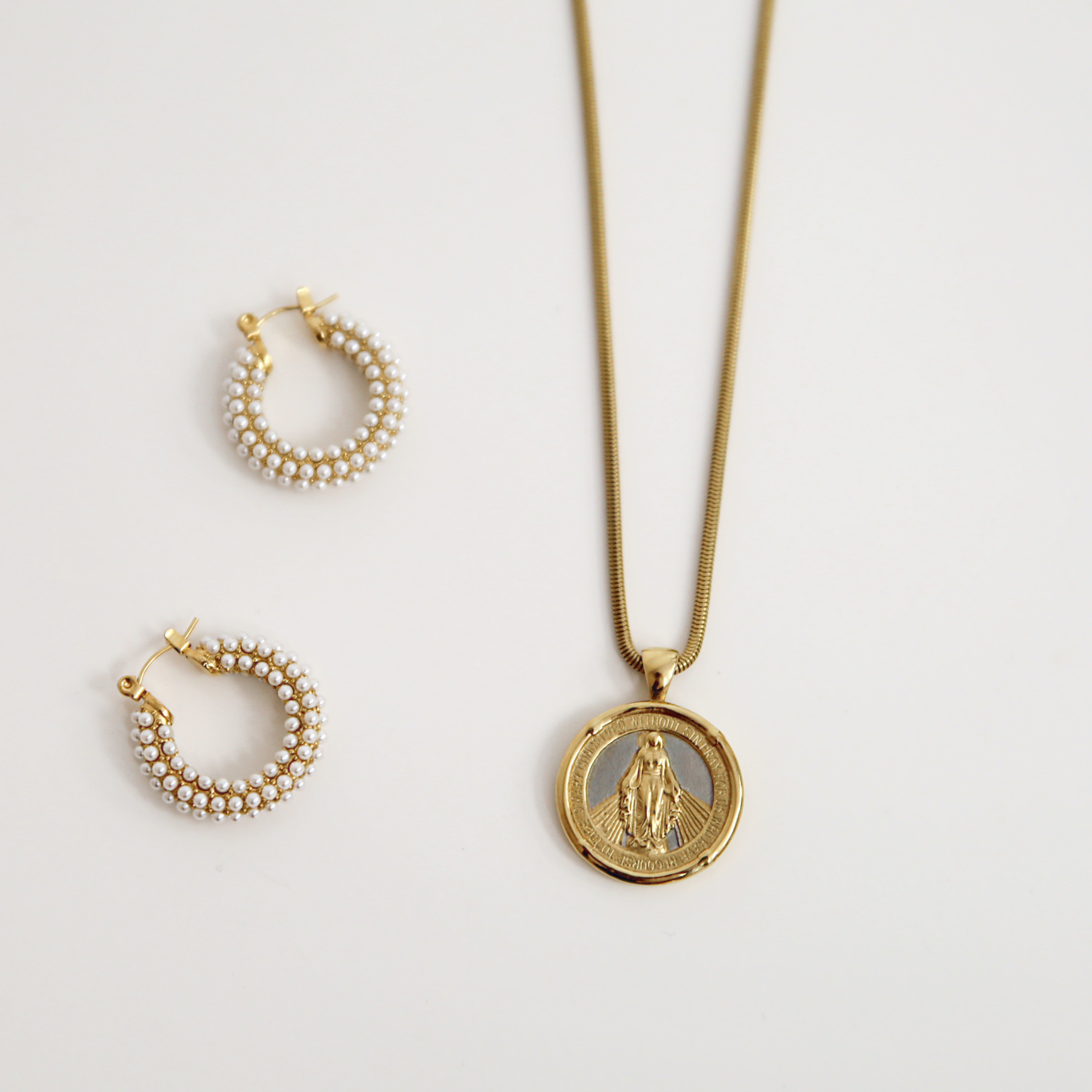 Gold necklace with a pendant and pearl hoop earrings on a light gray background