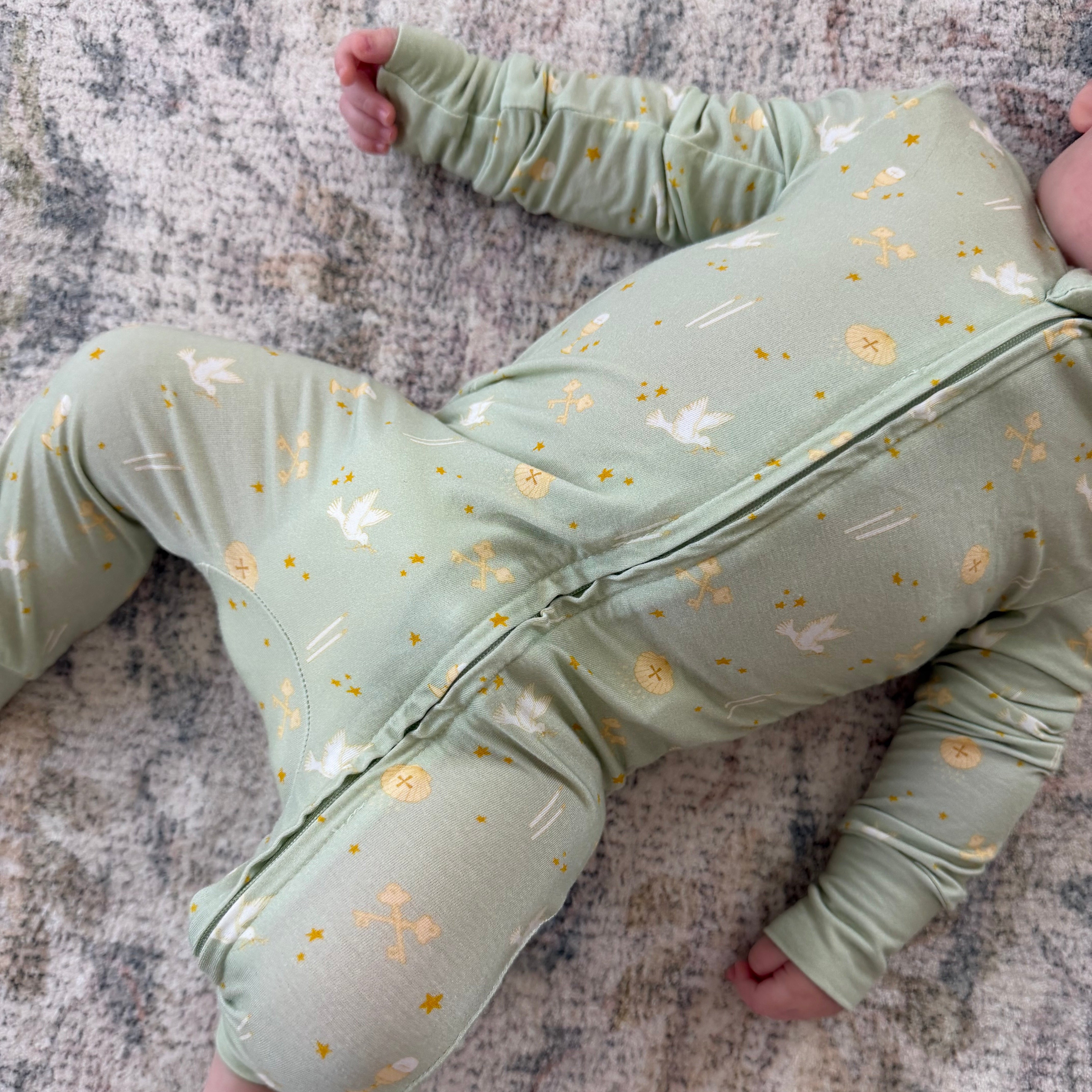 A baby lying on a textured gray rug wearing a sage green one-piece sleeper with a gold and white bunny print and a front zipper.